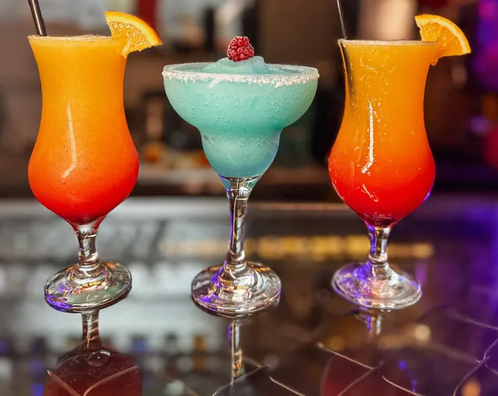 Três drinks no balcão: dois alaranjados com rodela de laranja e uma margarita azul com borda de açúcar