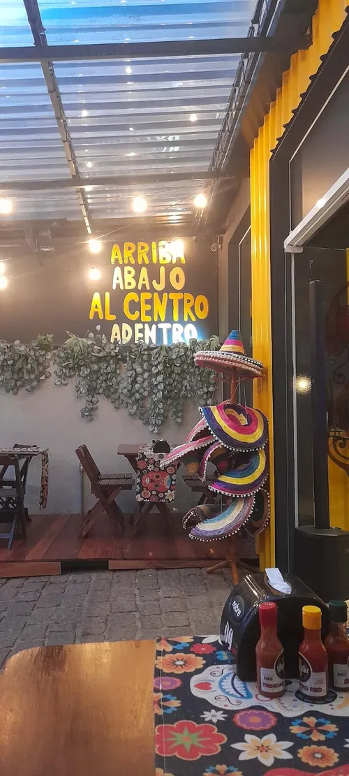 Parede da casa com o letreiro em neon Arriba, abajo, al centro, adentro, folhagens e sombreros coloridos